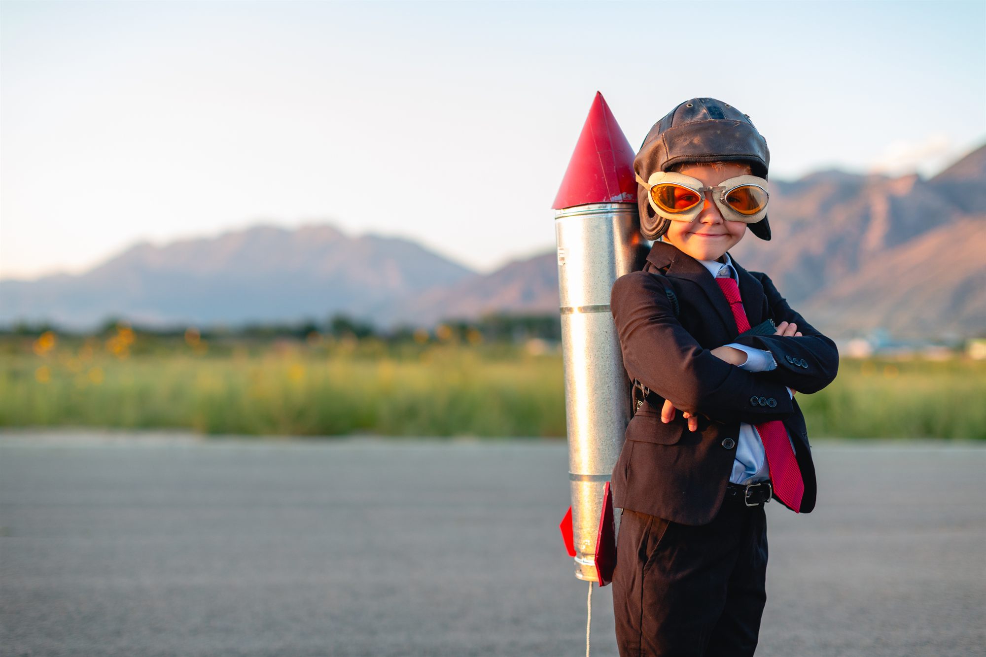 Kind im Anzug mit Fliegerbrille und selbstgebauter Rakete vor einer Bergkulisse — Sinnbild für große Ambitionen.